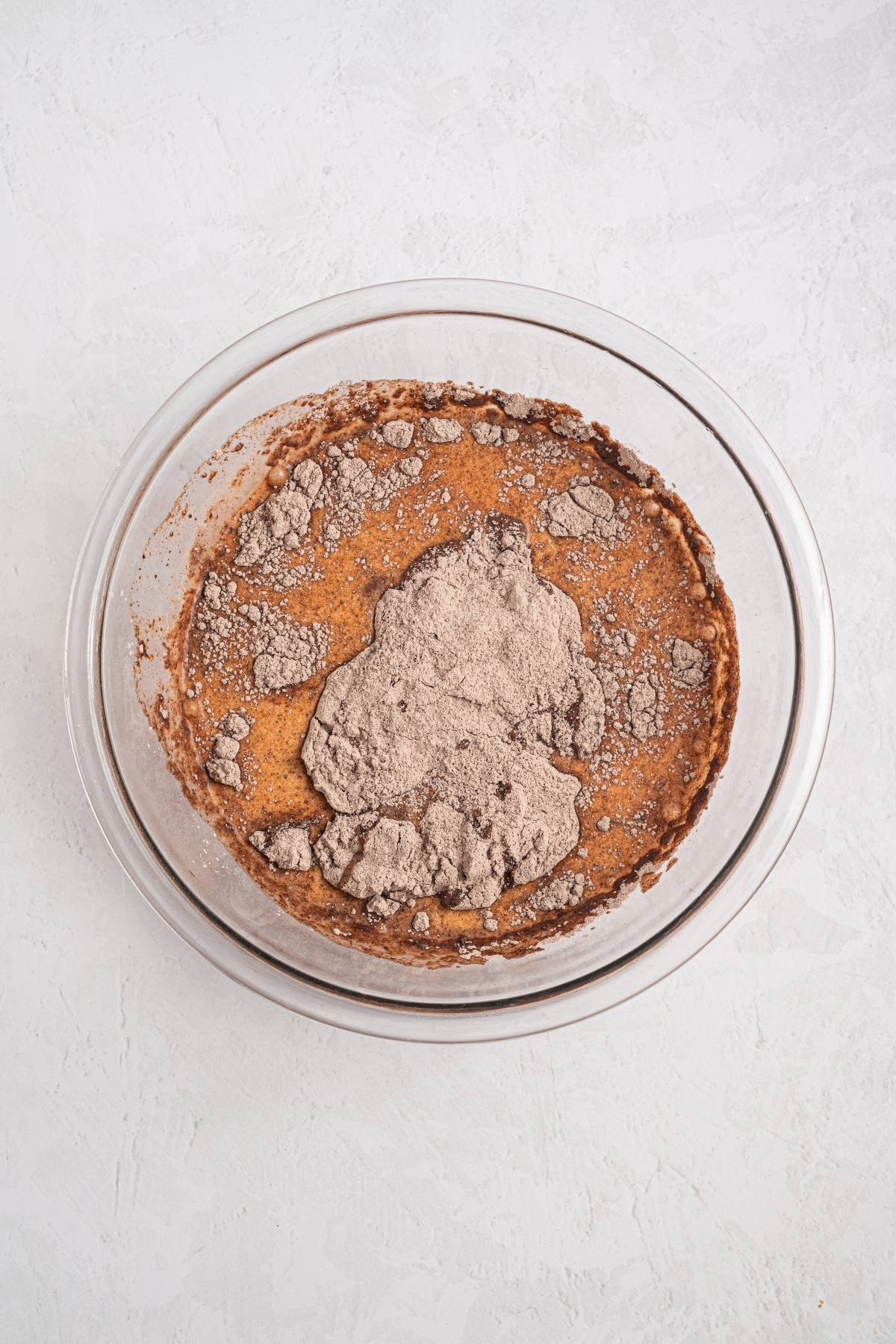 A glass bowl with brown batter and dry cocoa powder on top, set on a light textured surface.