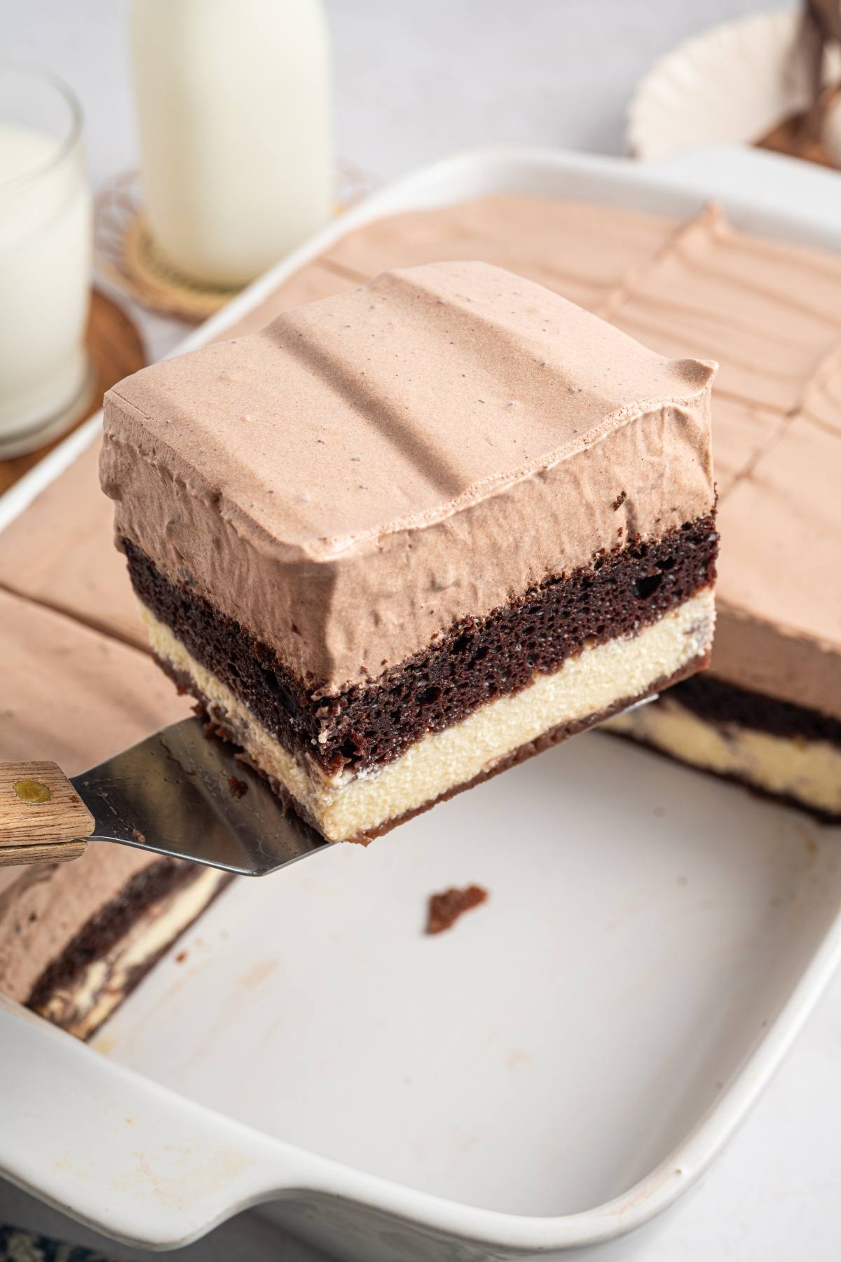 A spatula lifts a layered chocolate and vanilla cake slice with chocolate frosting from a baking dish.