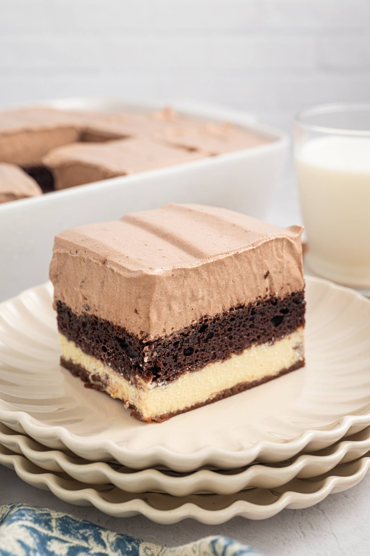 A slice of layered chocolate and vanilla cake with chocolate frosting on a plate, with milk in the background.