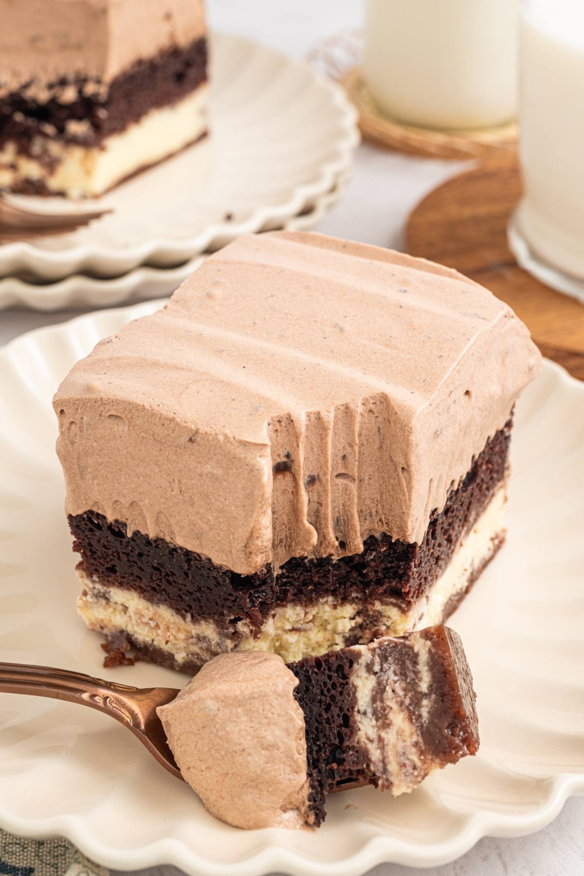 A slice of layered chocolate and cream cake with a bite taken out, on a white plate with a fork.