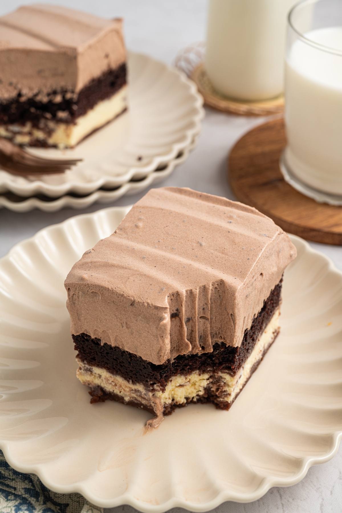 A slice of layered chocolate and cream cake with a bite taken out, on a cream-colored plate.