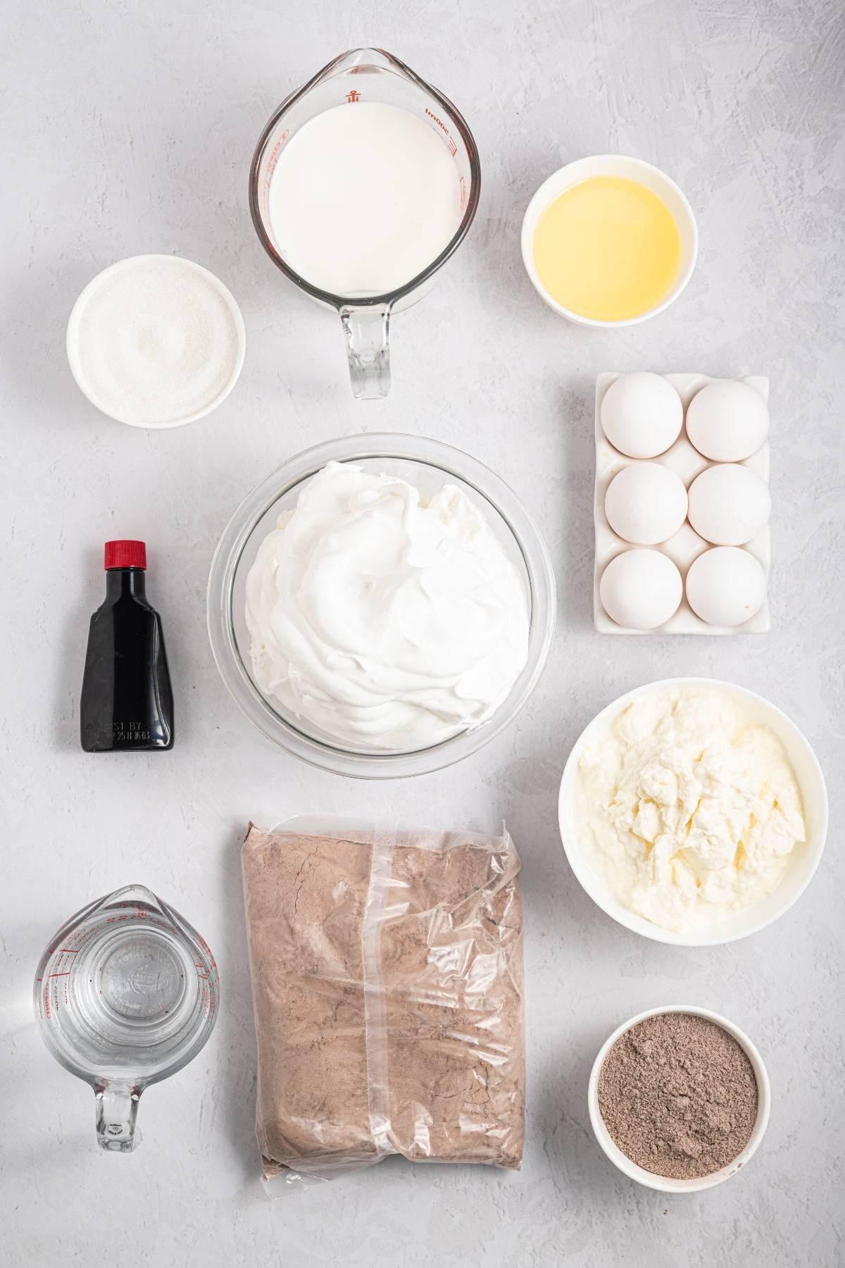 Top-down view of baking ingredients: eggs, whipped cream, cocoa mix, vanilla, milk, oil, sugar, and water.