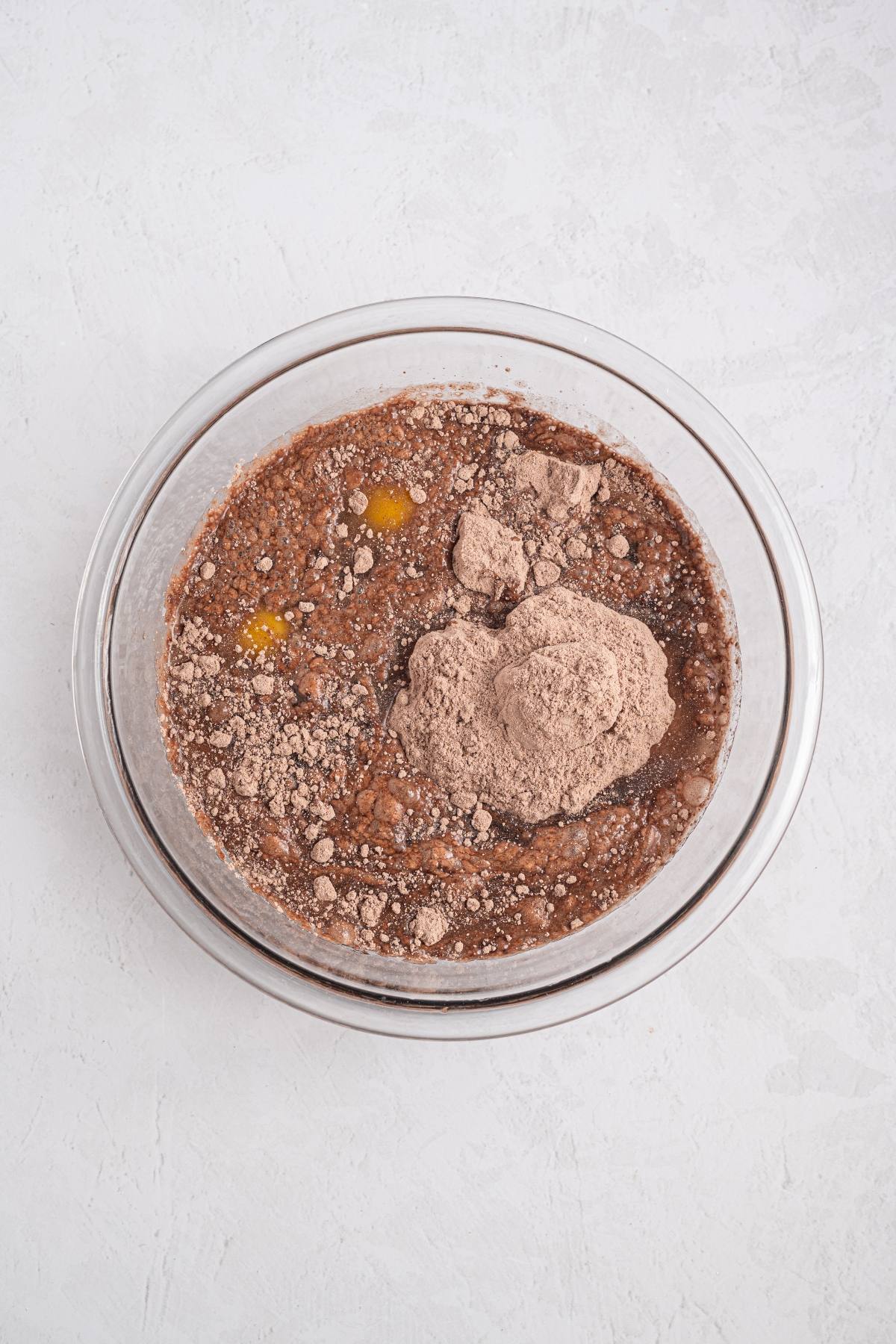 A glass bowl with brownie mix, eggs, and cocoa powder on a light textured surface.