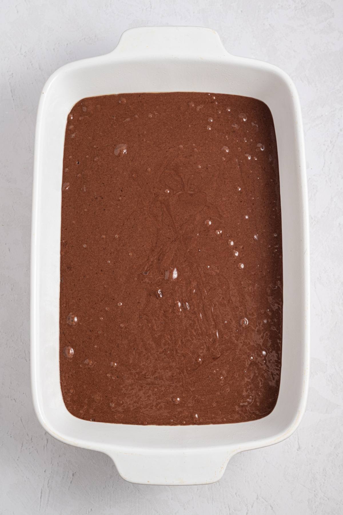 A white baking dish filled with chocolate cake batter on a light background.