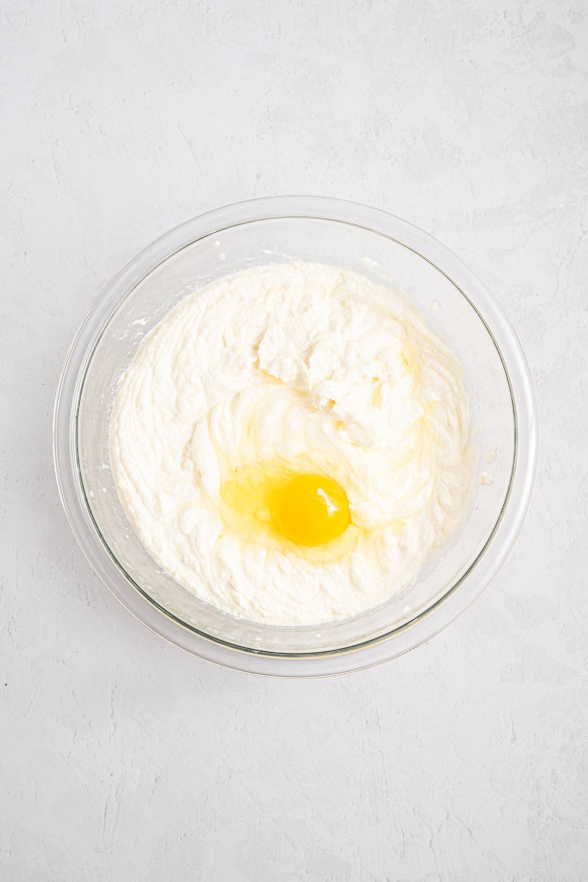 A glass bowl with creamy batter and a raw egg on top, on a light-colored surface.