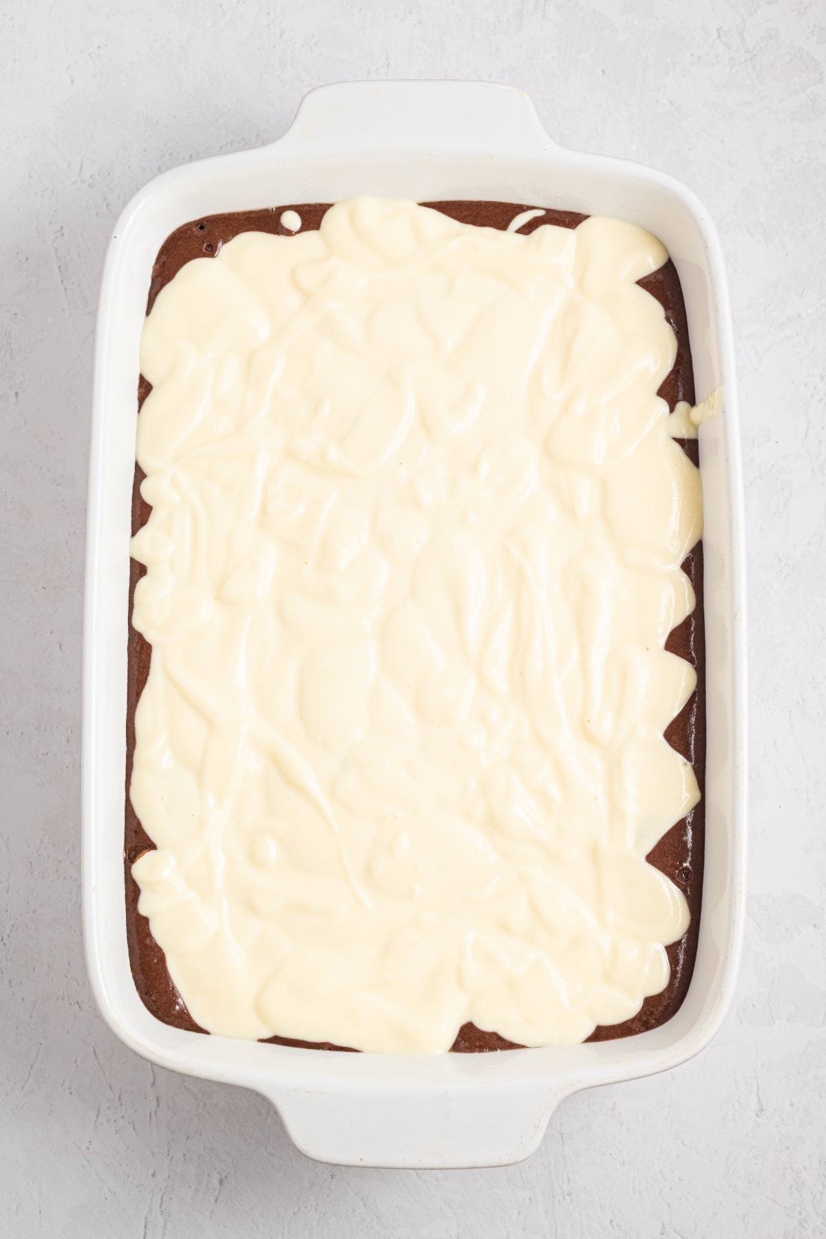 Rectangular baking dish with dark cake topped with a layer of creamy white batter or frosting.