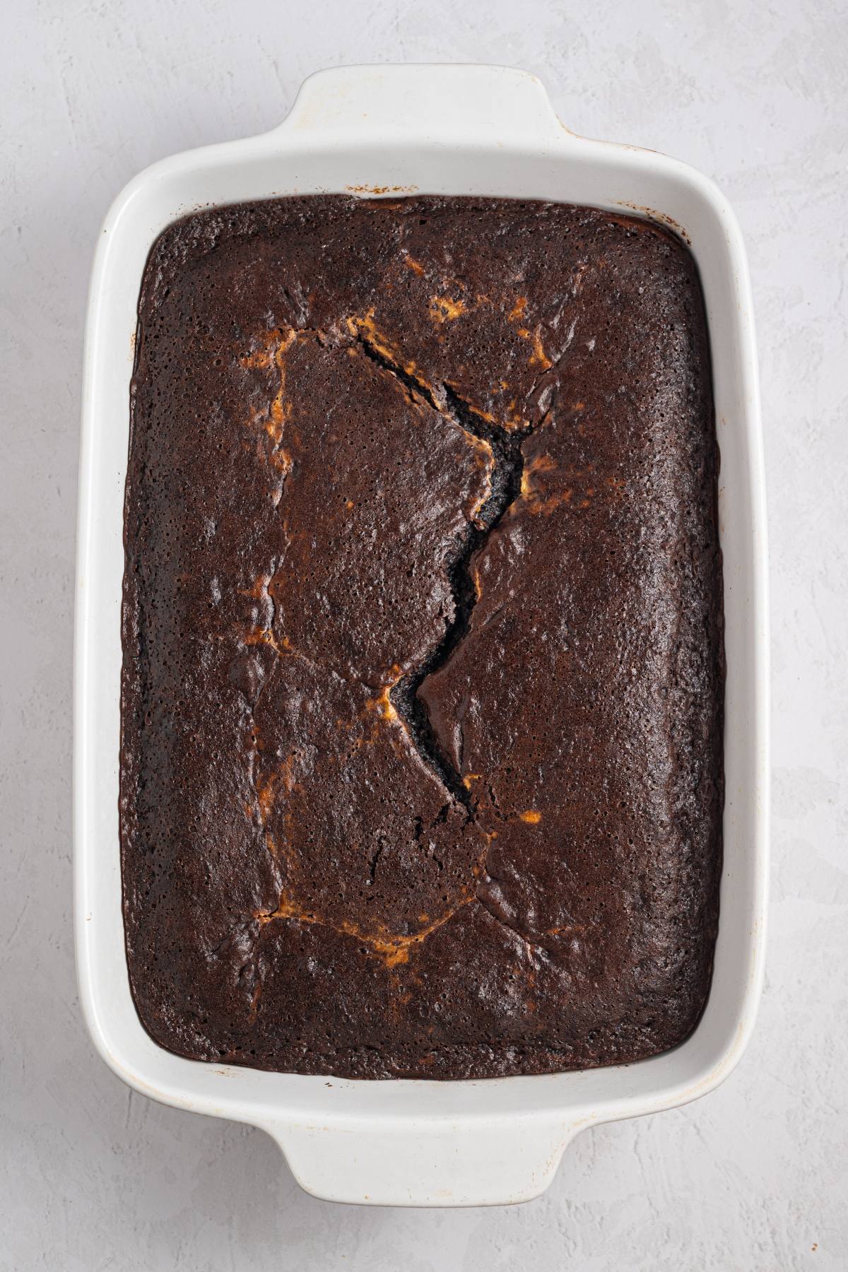 Rectangular chocolate cake with a cracked top in a white baking dish on a light surface.