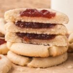 A stack of Jam Thumbprint Cookies filled with vibrant red jam sits among more cookies on a parchment surface.