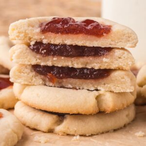 A stack of Jam Thumbprint Cookies filled with vibrant red jam sits among more cookies on a parchment surface.