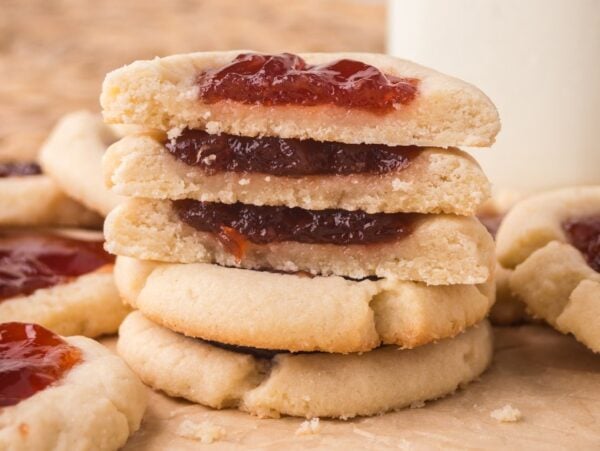 A stack of Jam Thumbprint Cookies filled with vibrant red jam sits among more cookies on a parchment surface.