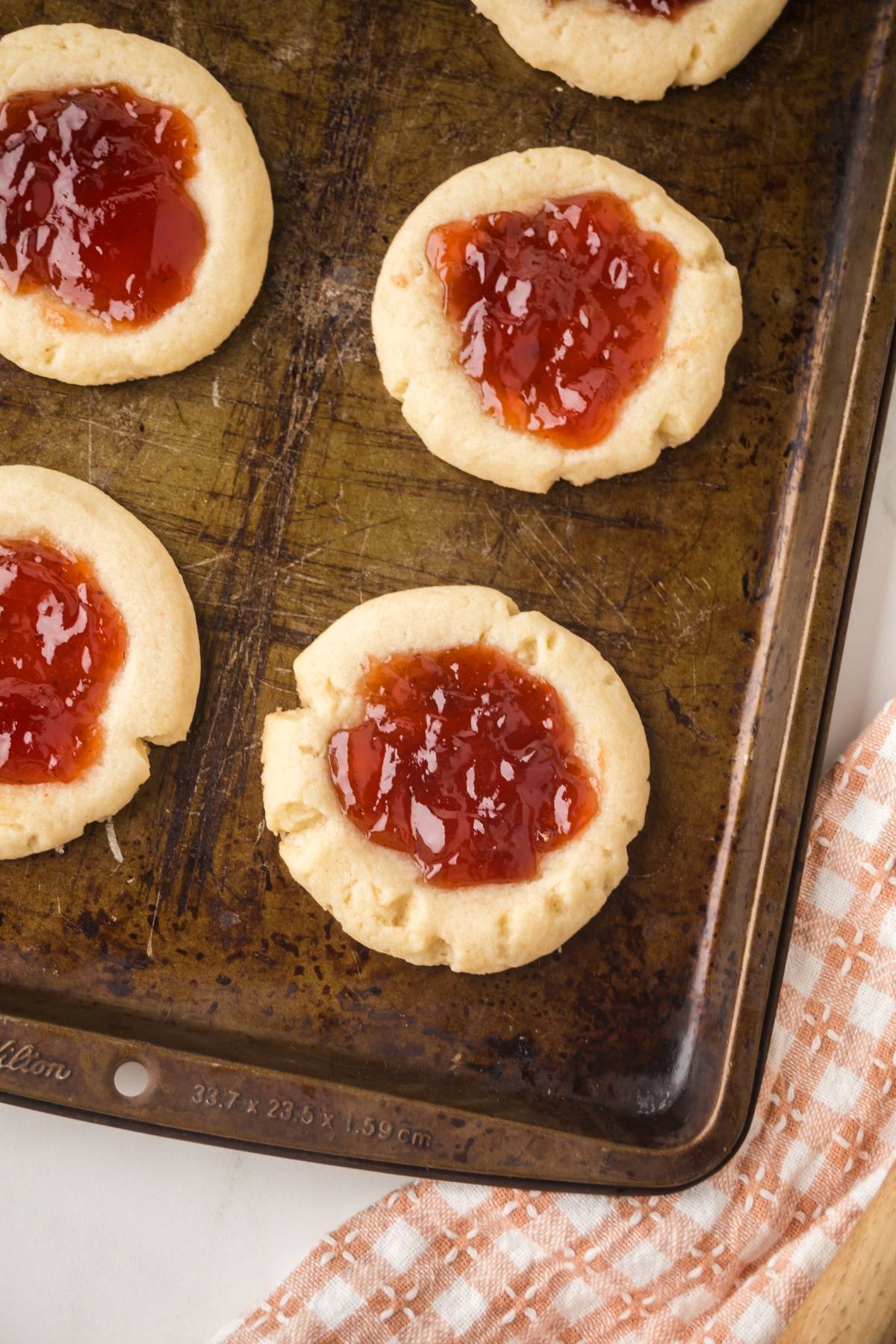 A baking sheet with thumbprint cookies filled with red jam, next to a checkered kitchen towel.