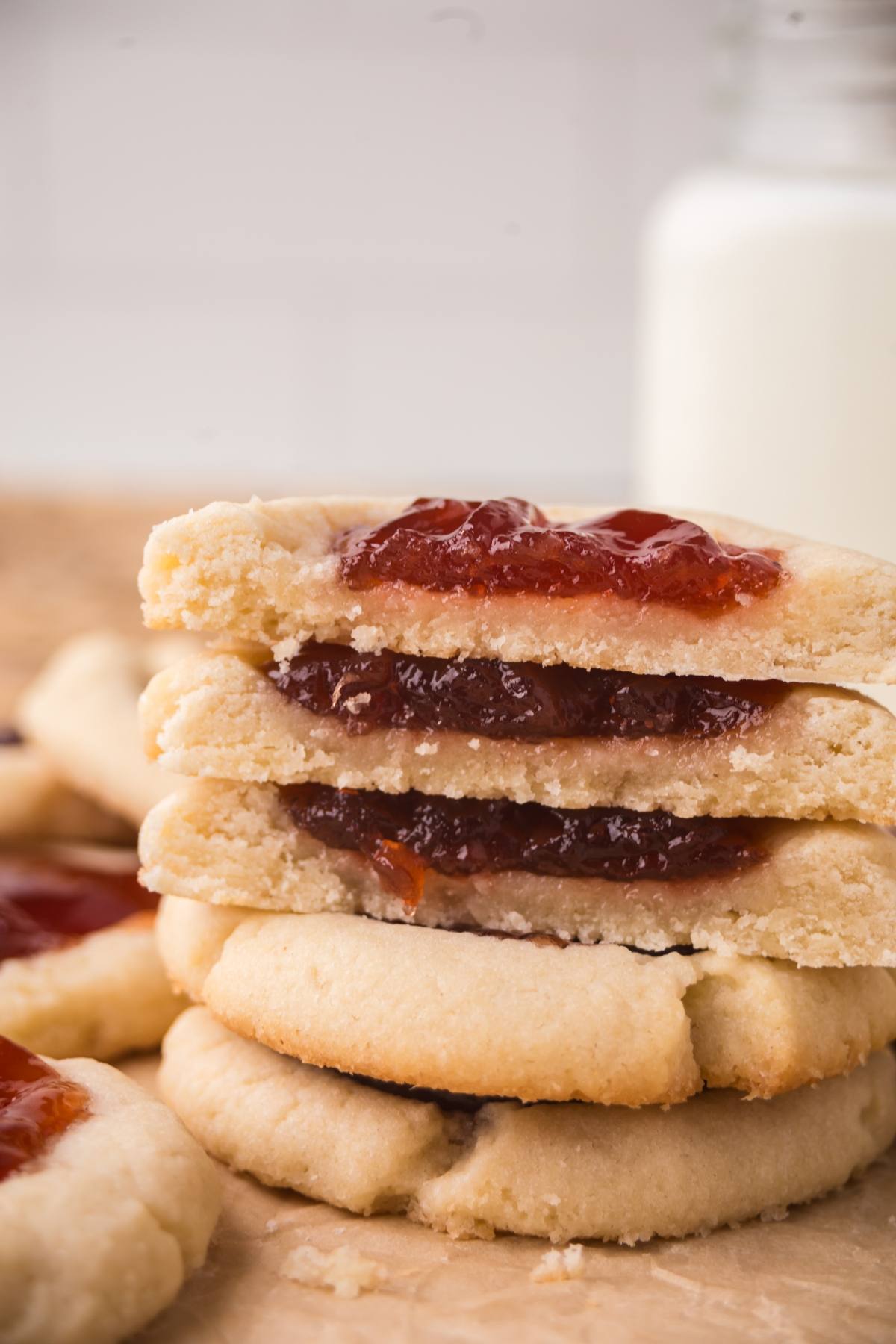 Stack of thumbprint cookies filled with red jam, with a glass of milk in the background.
