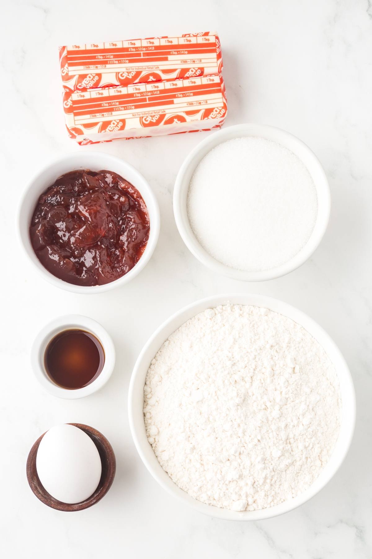 Bowl of flour, sugar, jam, an egg, vanilla extract, butter, and a small bowl of salt on a white surface.