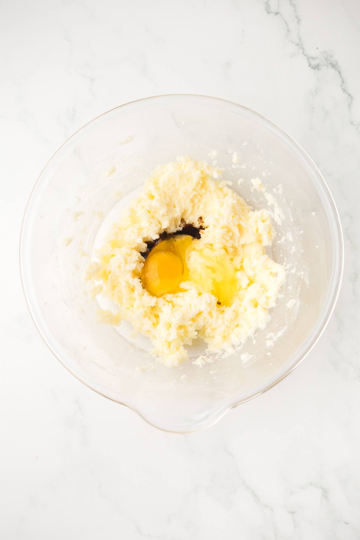 A glass bowl with butter, sugar, an egg, and vanilla being mixed on a white marble surface.