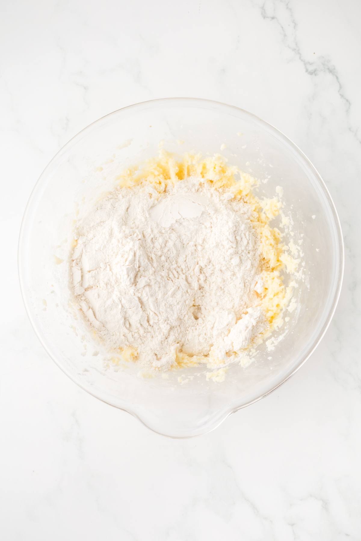 A glass bowl with flour and creamed butter and sugar on a white marble surface.