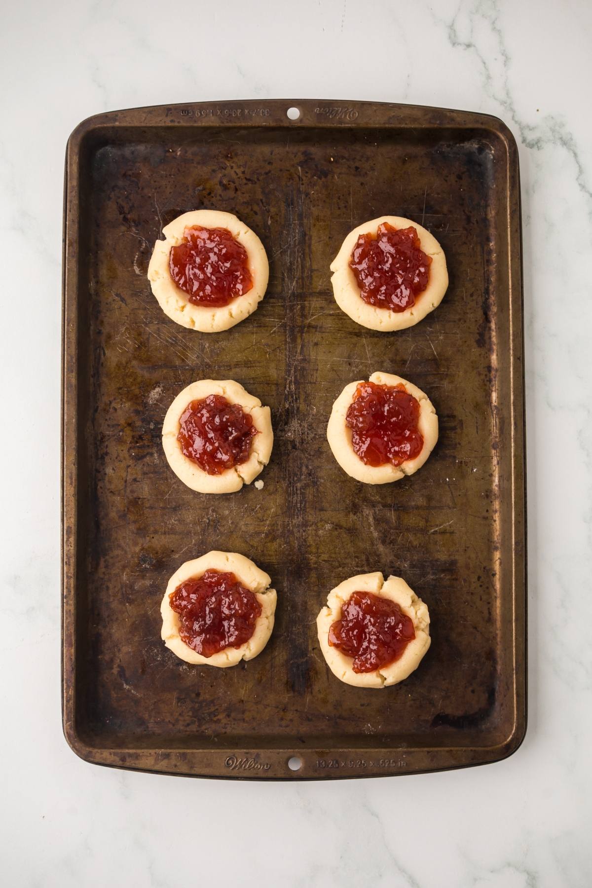 Six unbaked thumbprint cookies with red jam filling on a worn metal baking sheet.