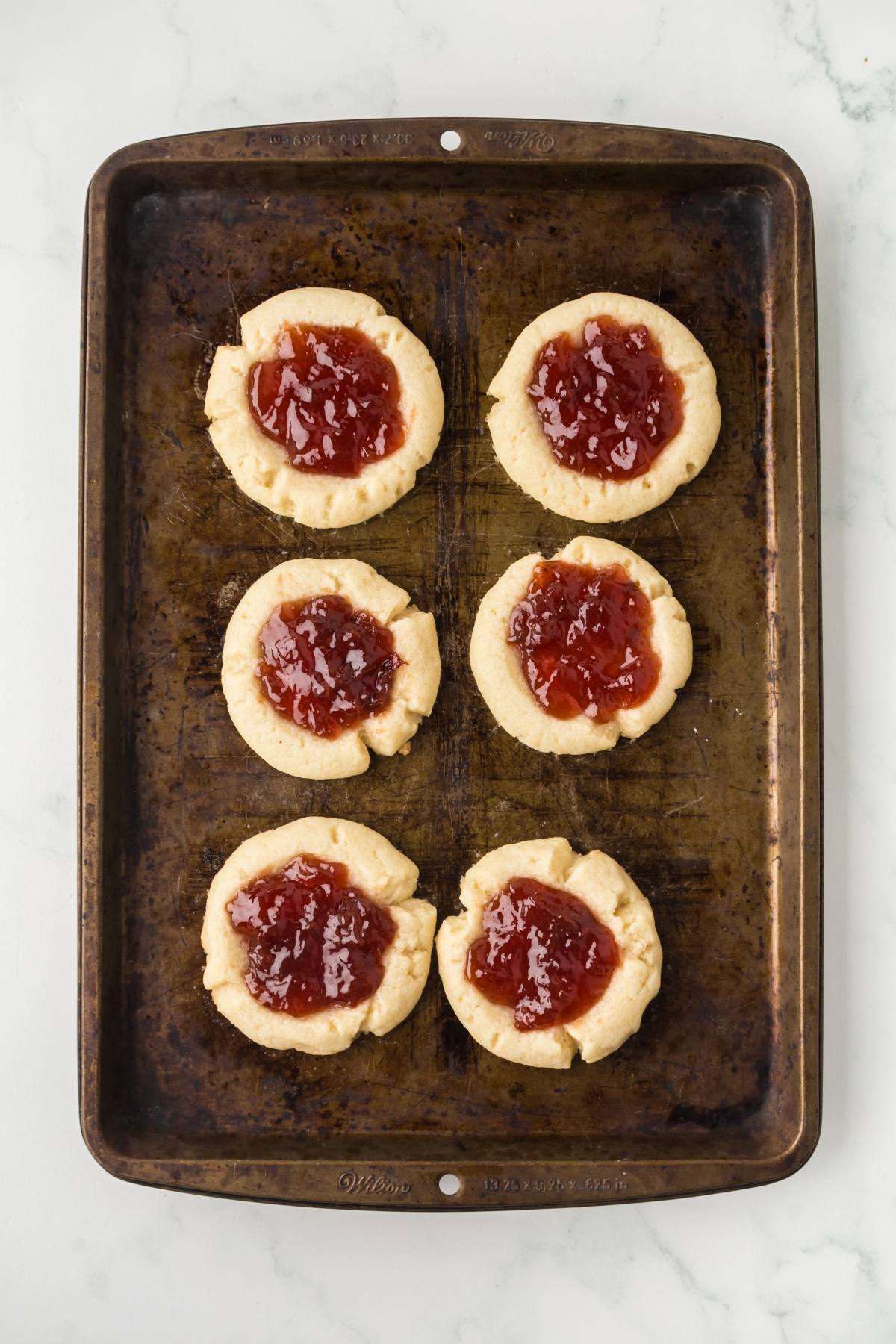 Six thumbprint cookies with red jam centers on a worn, rectangular baking sheet.