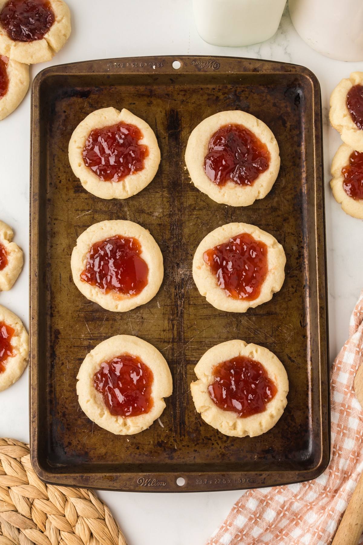 A baking sheet with six jam thumbprint cookies, surrounded by more cookies on a white surface.