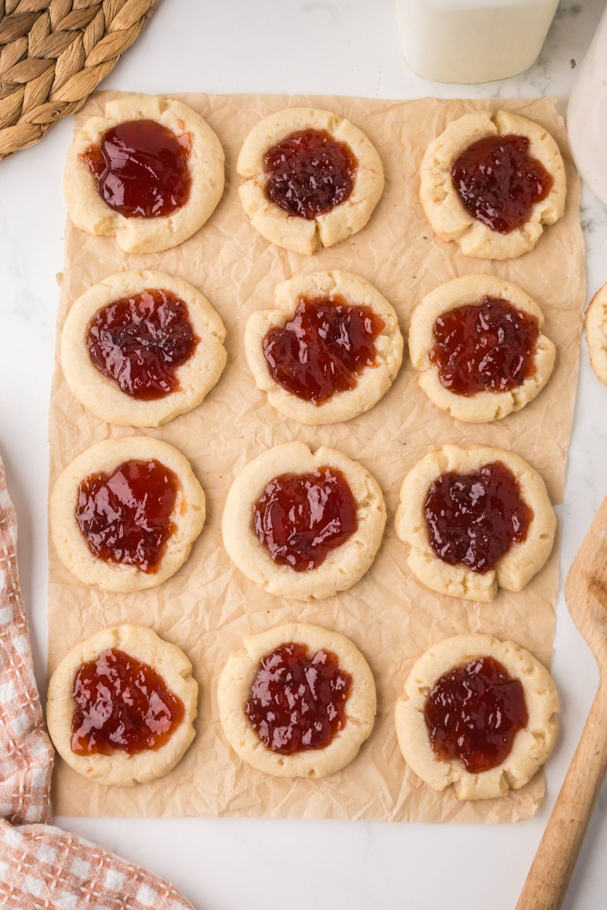 Twelve jam-filled thumbprint cookies on parchment paper, arranged in a grid, with a jar and cloth nearby.