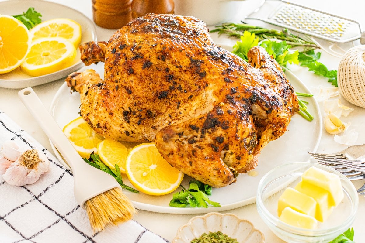 Whole lemon herb roasted chicken on a plate with lemon slices, fresh herbs, garlic, and butter arranged on the table.