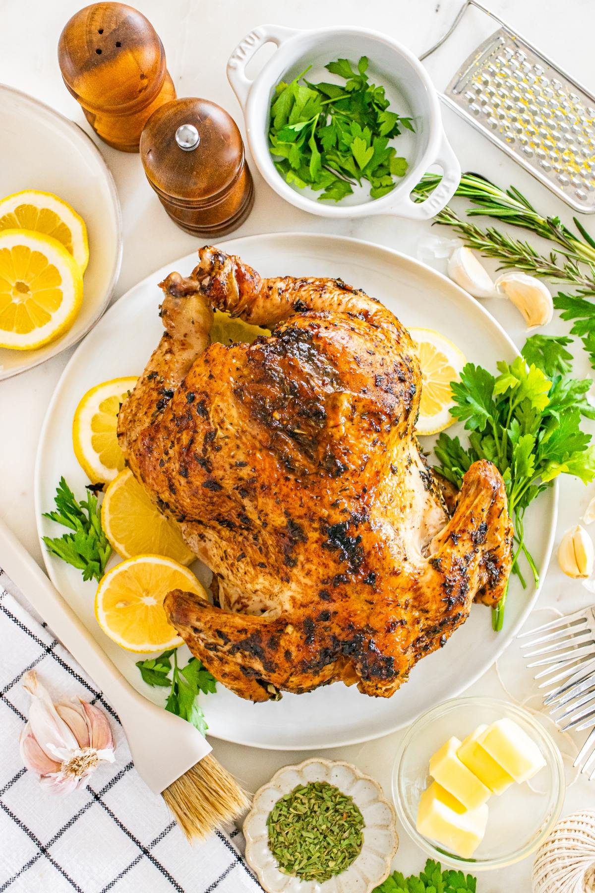 Roast chicken on a platter with lemon slices, fresh herbs, and seasoning ingredients arranged around it.
