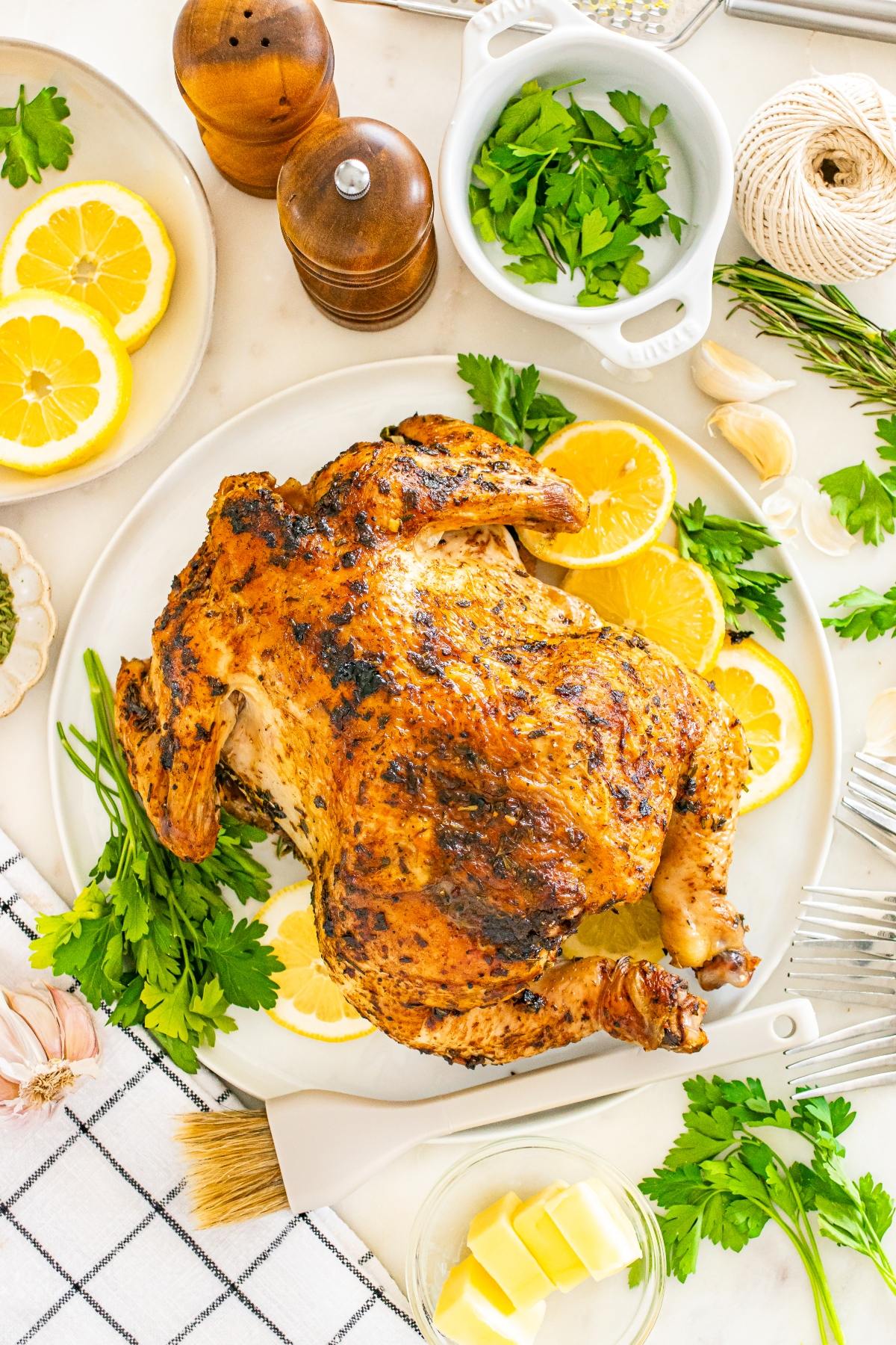 Roast chicken on a plate with lemon slices, parsley, and spices, surrounded by herbs and kitchen items.