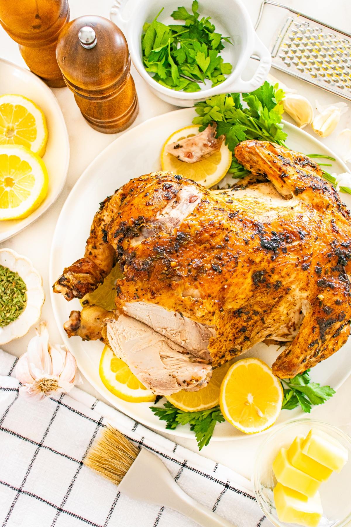 Roast chicken on a plate with lemon slices, parsley, butter, and seasonings on a white table.