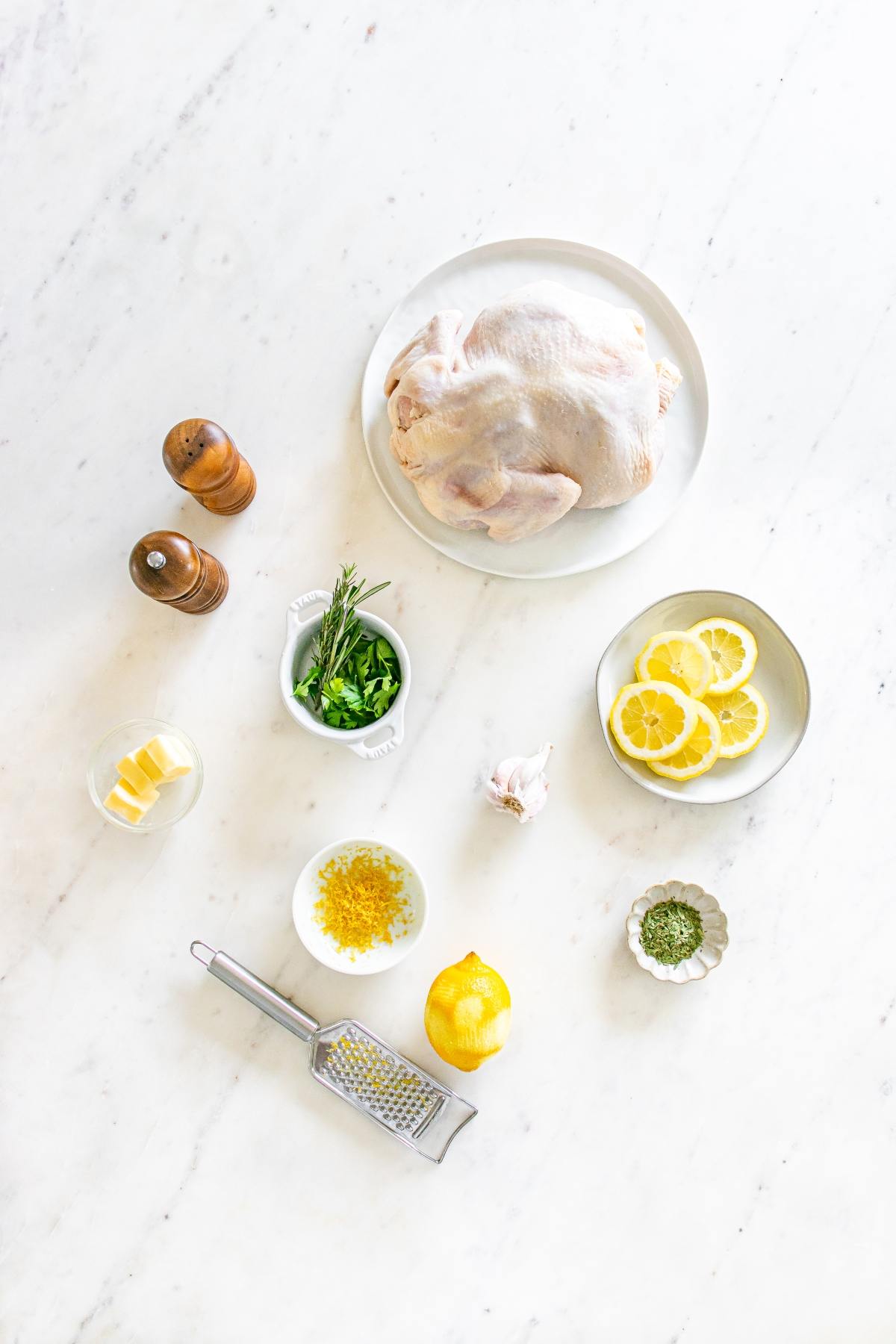 Ingredients for roast chicken: raw whole chicken, lemon, lemon slices, herbs, butter, garlic, salt, and pepper.