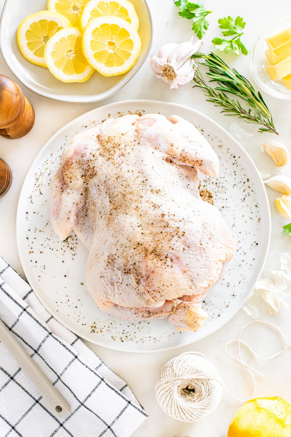 Raw whole chicken seasoned with pepper on a white plate, surrounded by lemon slices, herbs, garlic, and kitchen tools.