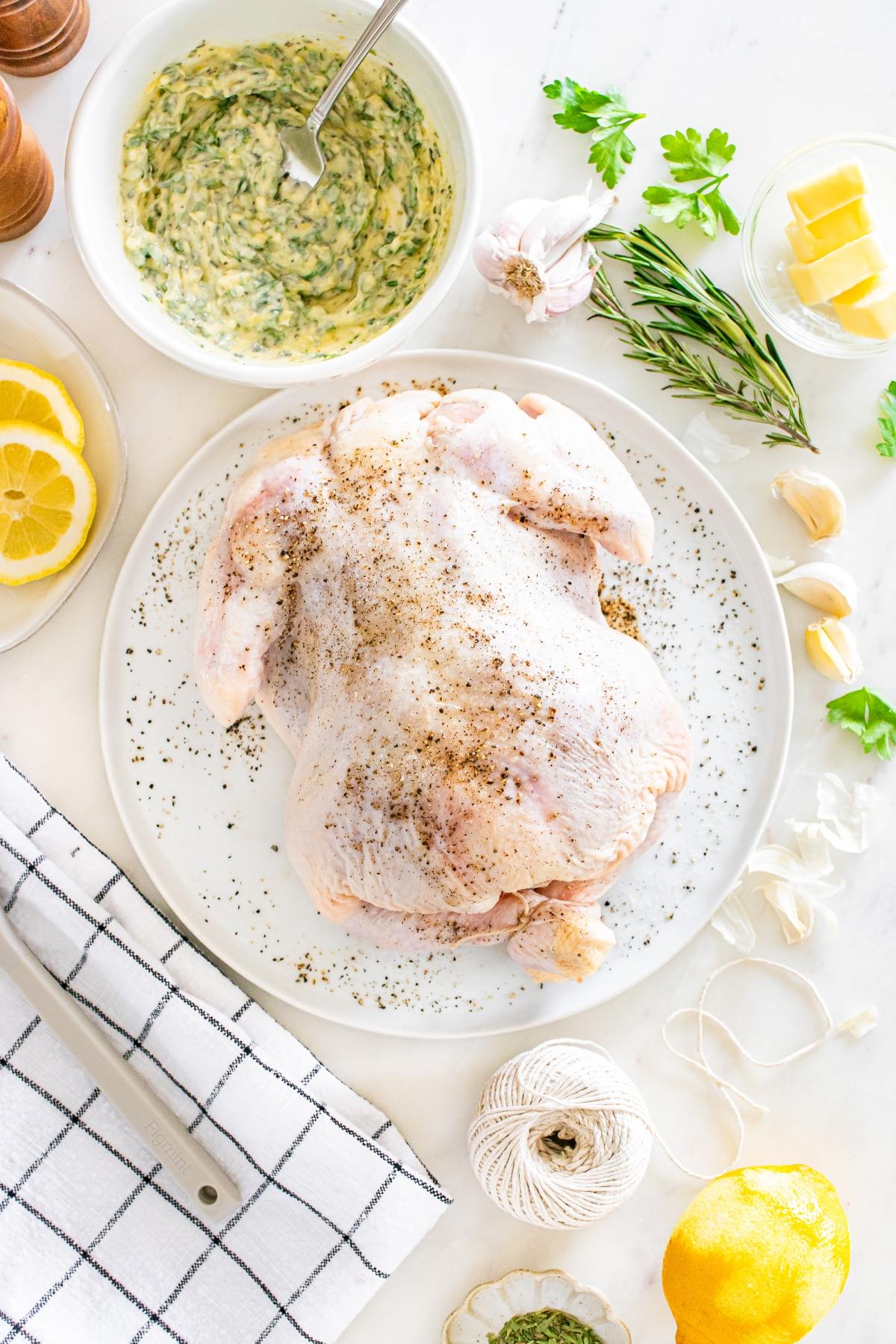 Raw whole chicken on a plate with herbs, garlic, lemon, butter, and seasoning on a white countertop.