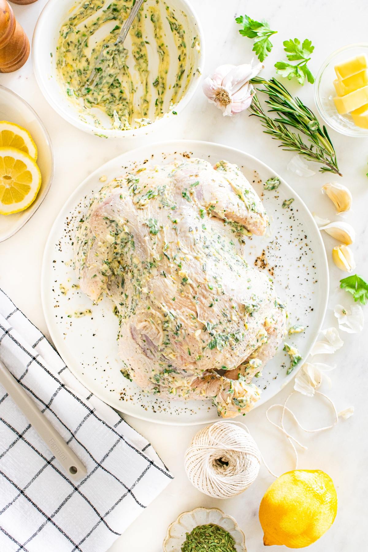 Raw whole chicken coated with herb butter on a plate, surrounded by lemon, herbs, garlic, and kitchen twine.