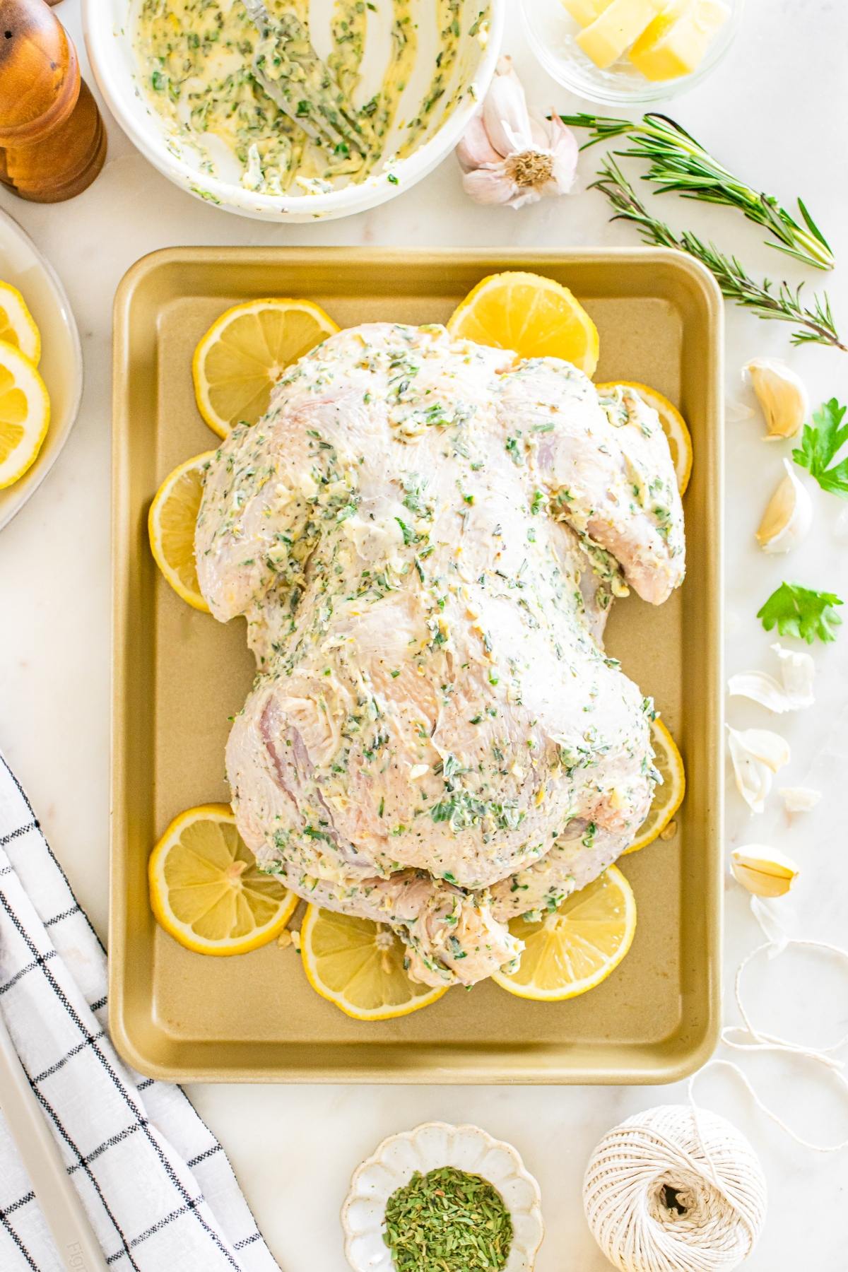 Whole raw chicken coated in herb butter on a baking sheet, surrounded by lemon slices and fresh herbs.