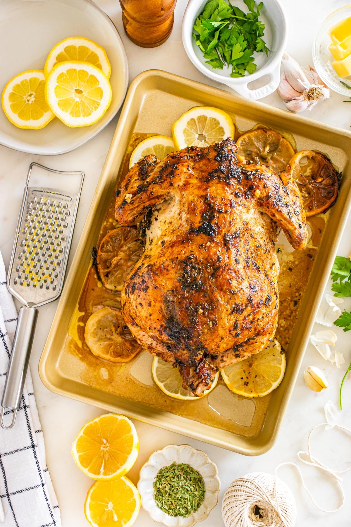 Roast chicken on a baking sheet with lemon slices, herbs, and kitchen utensils arranged around it.