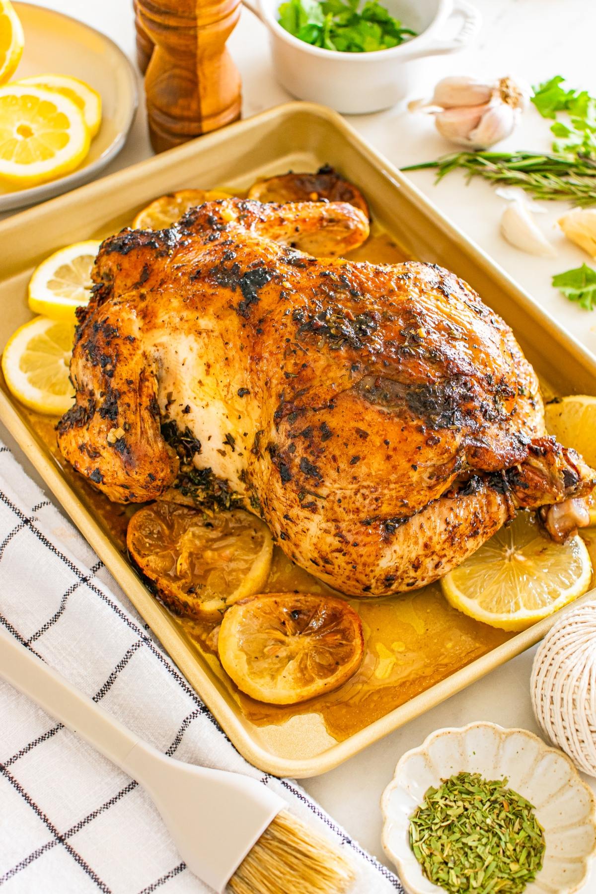 Roast chicken with herbs on a baking sheet, surrounded by lemon slices and fresh ingredients.