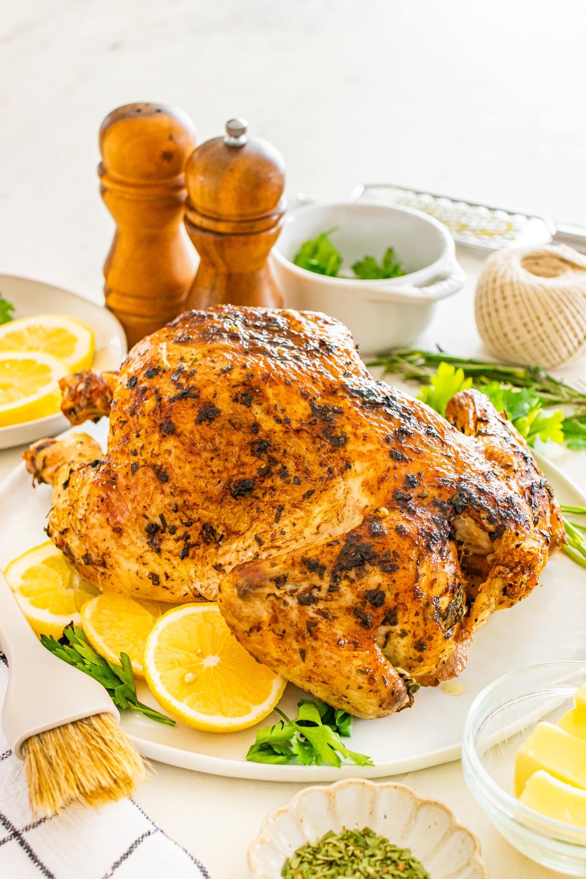 A whole roasted chicken on a platter with lemon slices and parsley, surrounded by spices and kitchen tools.