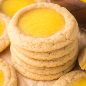 A stack of Lemon Thumbprint Cookies surrounded by more cookies sits on parchment paper, showcasing their delightful lemon curd centers.