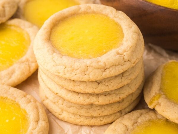 A stack of Lemon Thumbprint Cookies surrounded by more cookies sits on parchment paper, showcasing their delightful lemon curd centers.