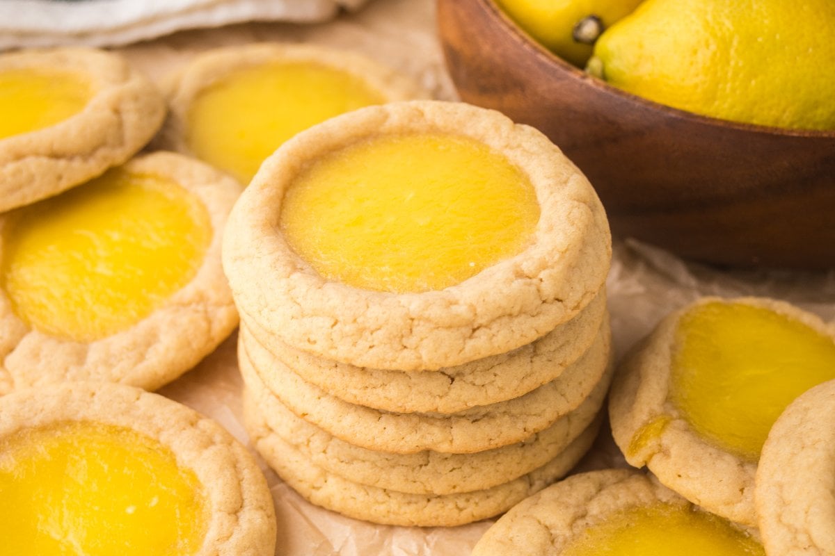 A stack of Lemon Thumbprint Cookies surrounded by more cookies and a bowl of fresh lemons.