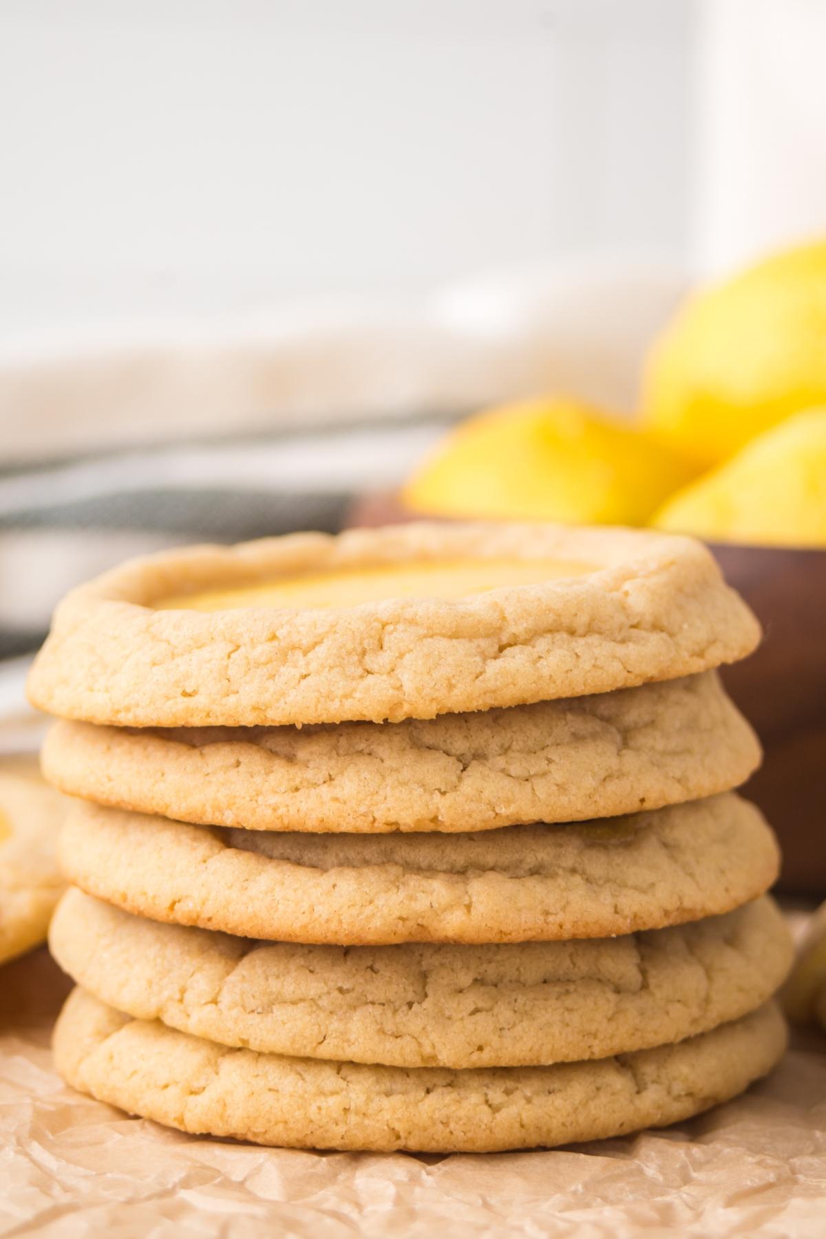 A stack of five round, golden Lemon Thumbprint cookies on parchment paper with lemons in the background.