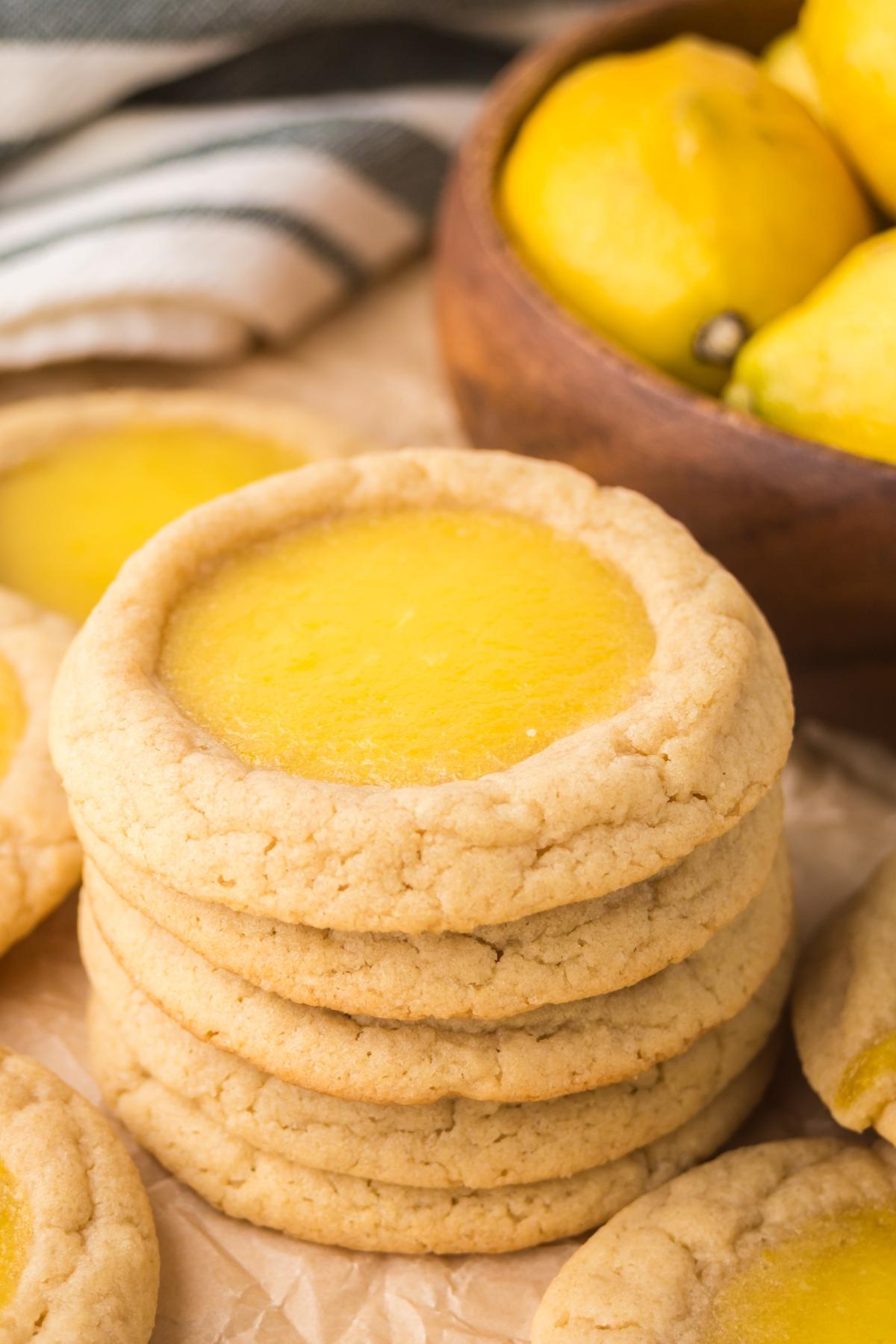 Stack of lemon Thumbprint cookies with yellow centers, with fresh lemons in a wooden bowl in the background.