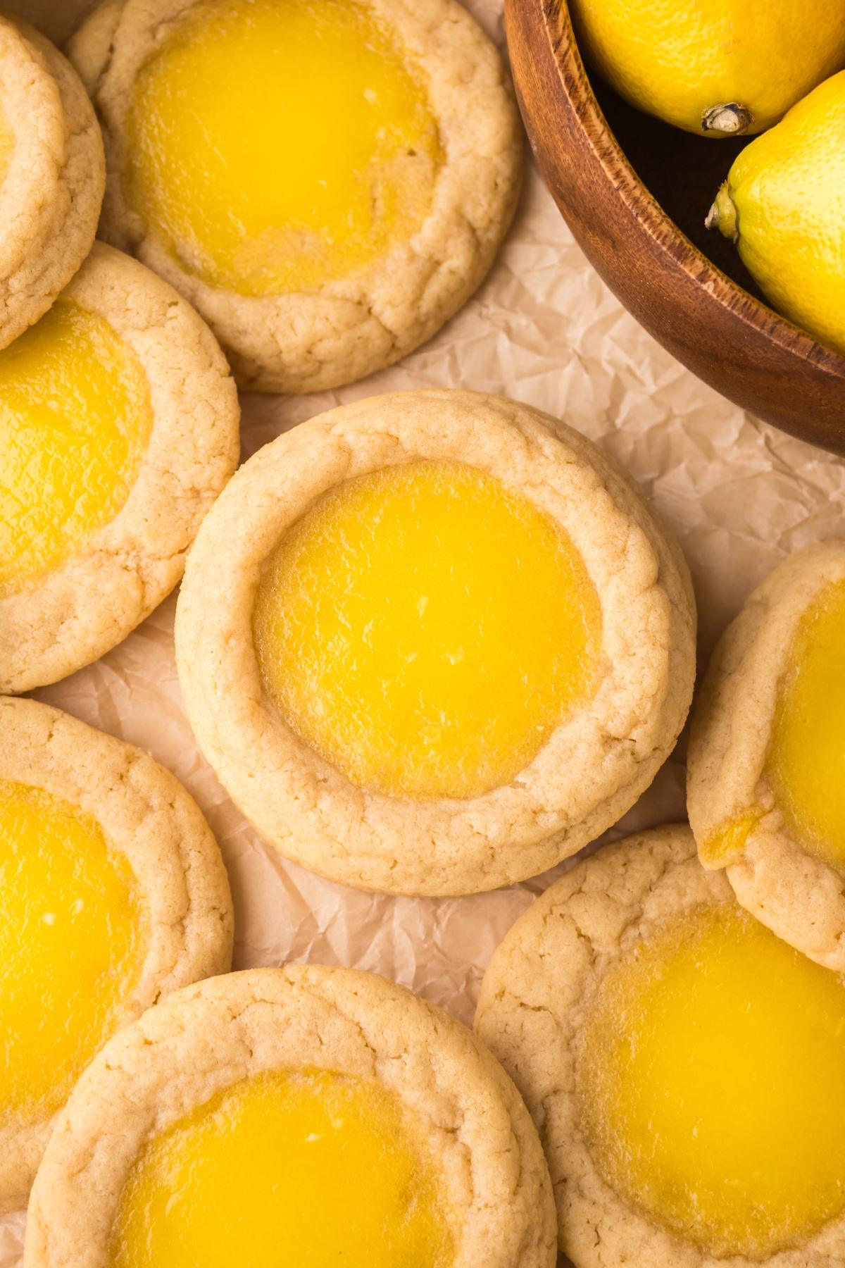 Lemon Thumbprint cookies with bright yellow centers arranged on parchment next to a wooden bowl of fresh lemons.