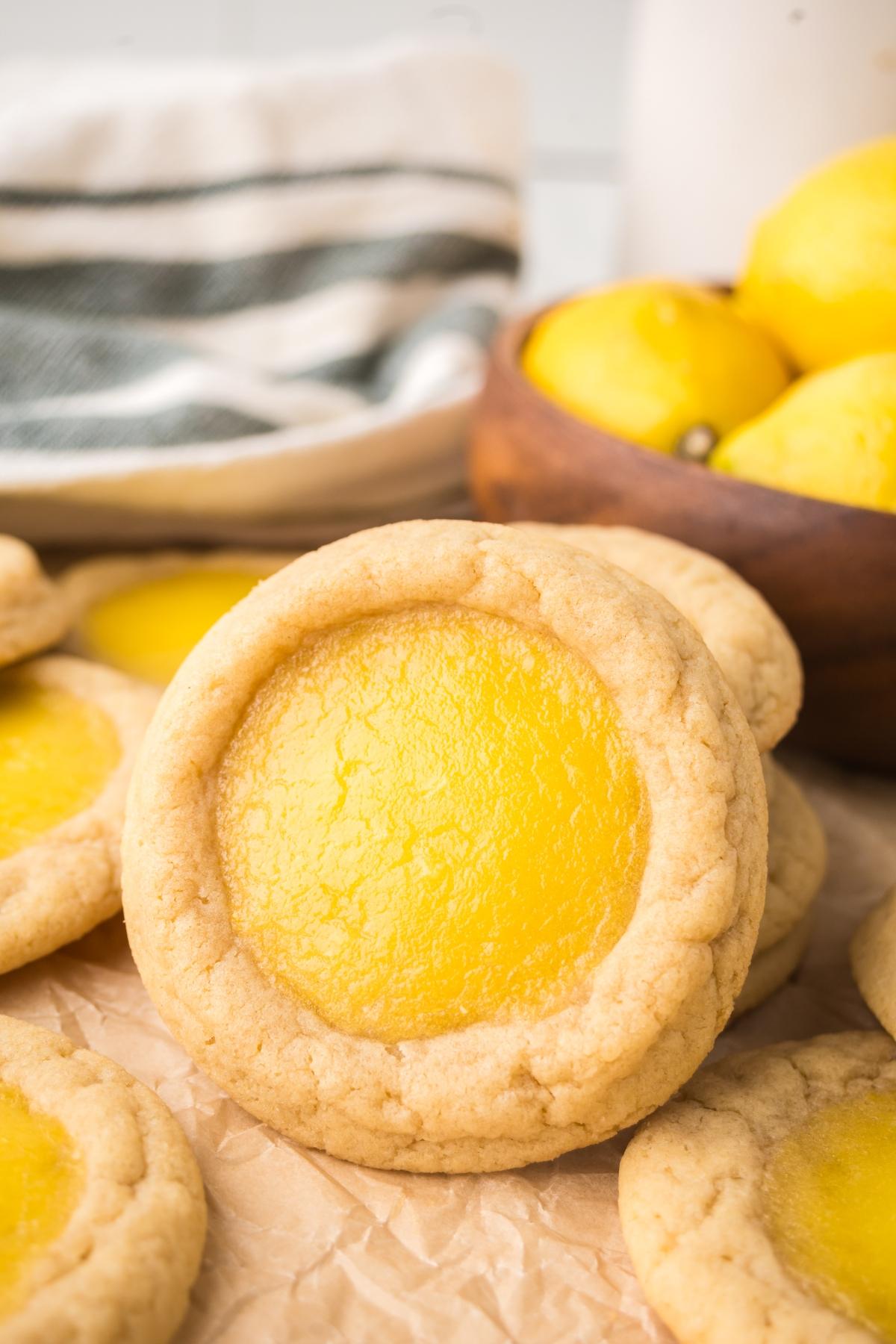 A close-up of a lemon thumbprint cookie with a yellow filling, surrounded by more cookies and fresh lemons.