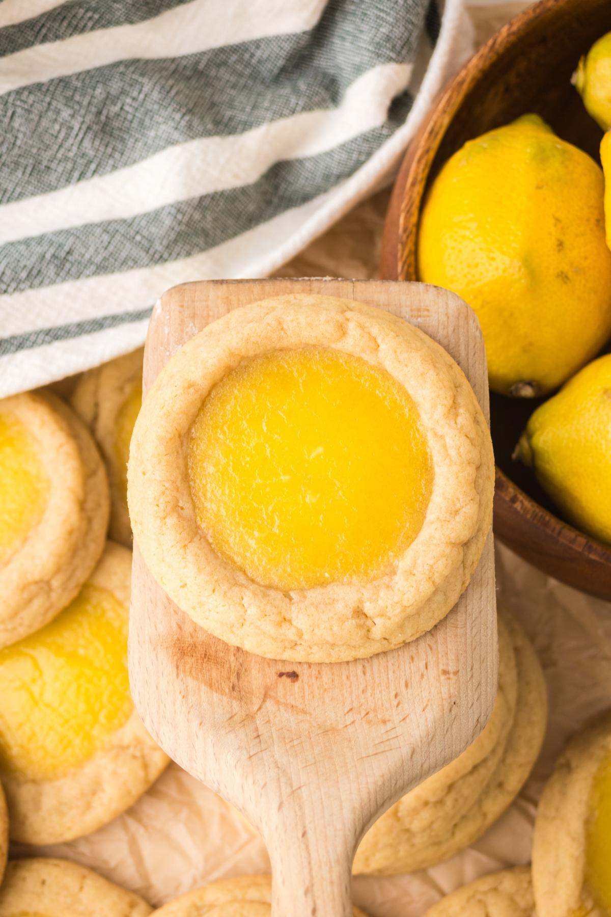 A lemon Thumbprint cookie with yellow filling on a wooden spatula, next to whole lemons and more cookies.