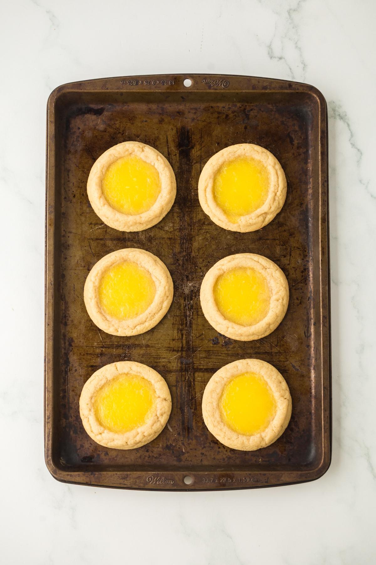 Six round lemon Thumbprint cookies on a dark baking sheet, placed on a white marble surface.