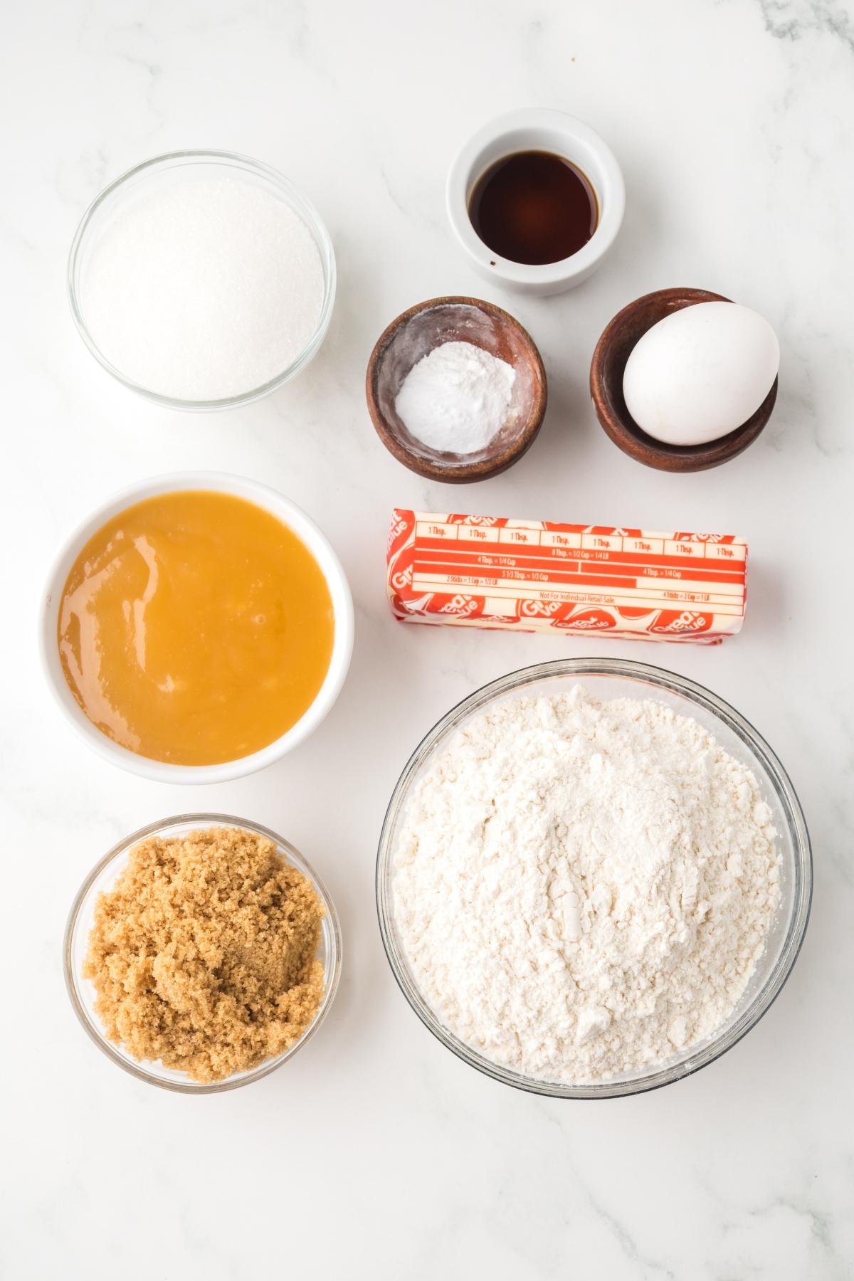 Baking ingredients on a white surface: flour, sugars, butter, egg, vanilla, honey, baking soda, and salt in bowls.