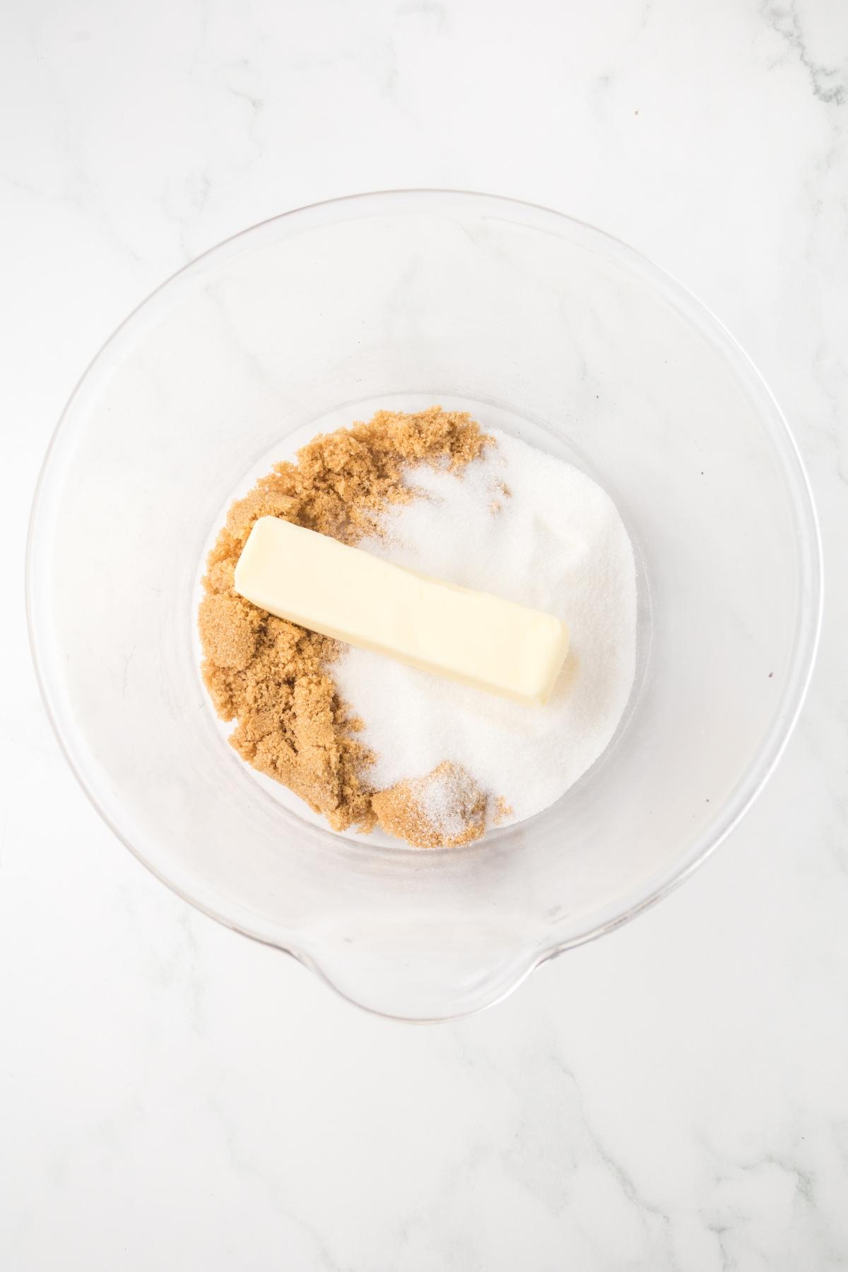A glass bowl with a stick of butter, brown sugar, and white sugar on a white surface.