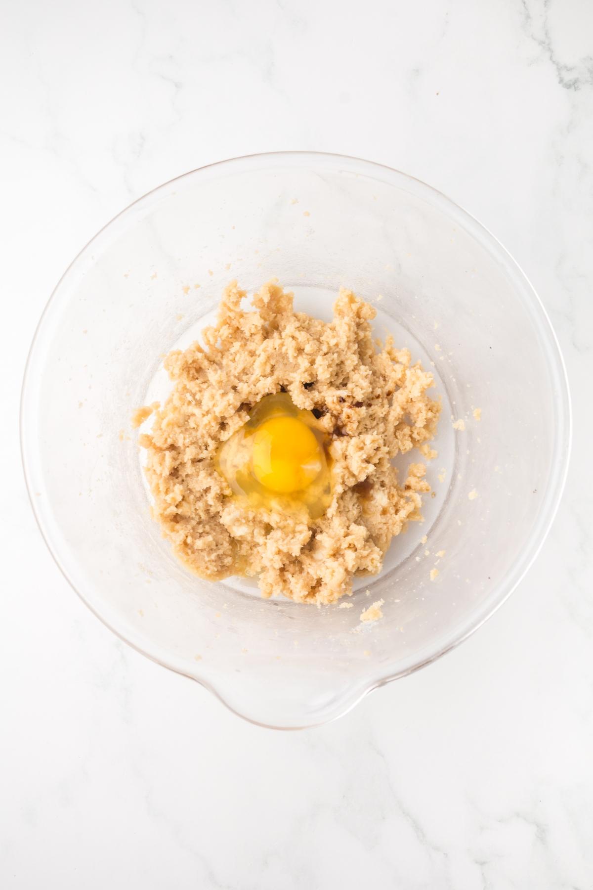 A glass bowl with cookie dough mixture and a raw egg in the center, on a white marble surface.