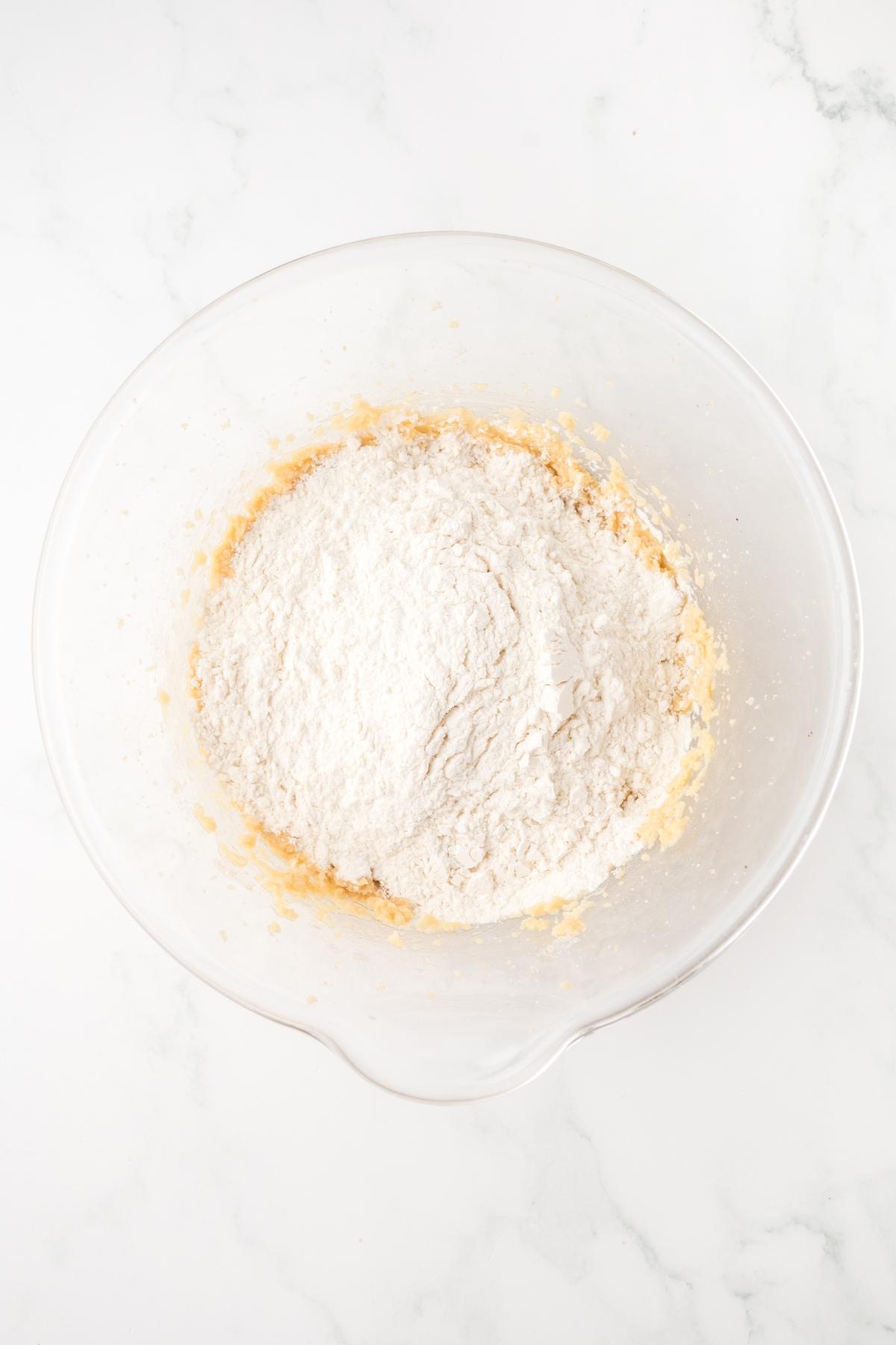 Clear mixing bowl with flour and cookie dough on a white marble surface, ready to be mixed.