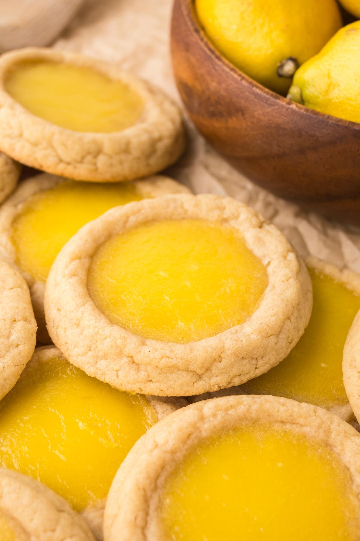 Lemon Thumbprint cookies on parchment paper next to a wooden bowl filled with fresh lemons.
