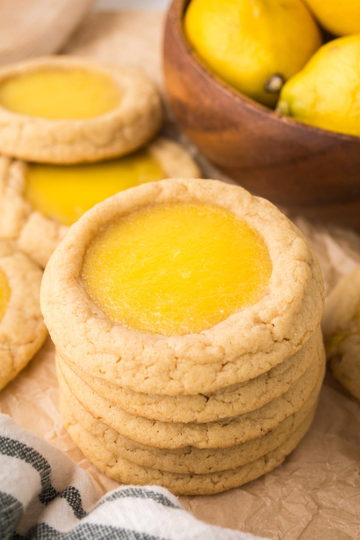 A stack of lemon Thumbprint cookies with yellow centers, next to a bowl of fresh lemons on a wooden surface.